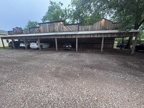 Ett överbyggt parkeringsområde med grusunderlag, träkonstruktion och fordon parkerade under taket.