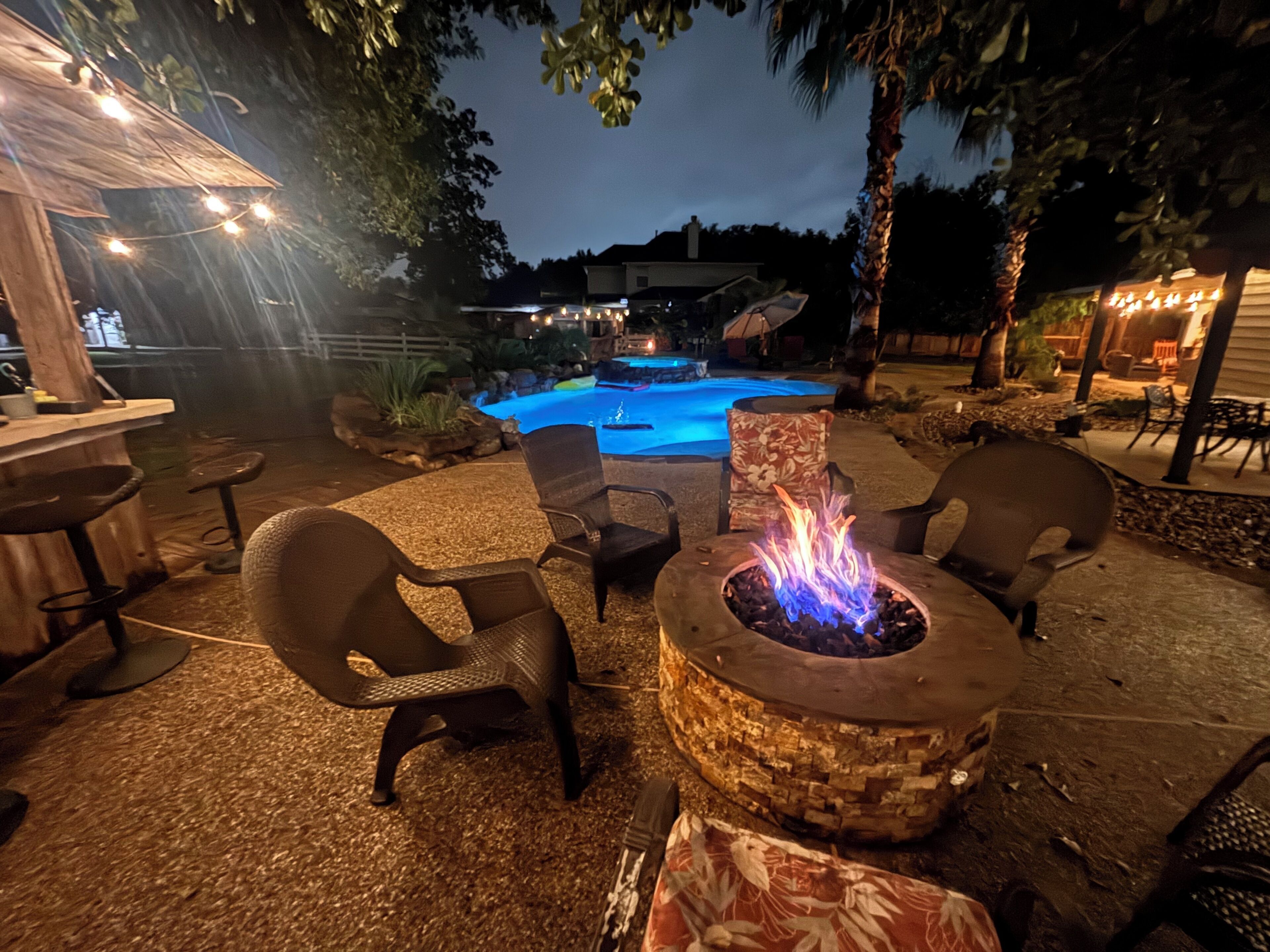 An outdoor patio area with a fire pit, seating, and a swimming pool in the background.