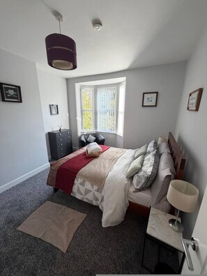 Ein Schlafzimmer mit Bett, Nachttisch, Lampe, Kommode, Fenster und Pendelleuchte.