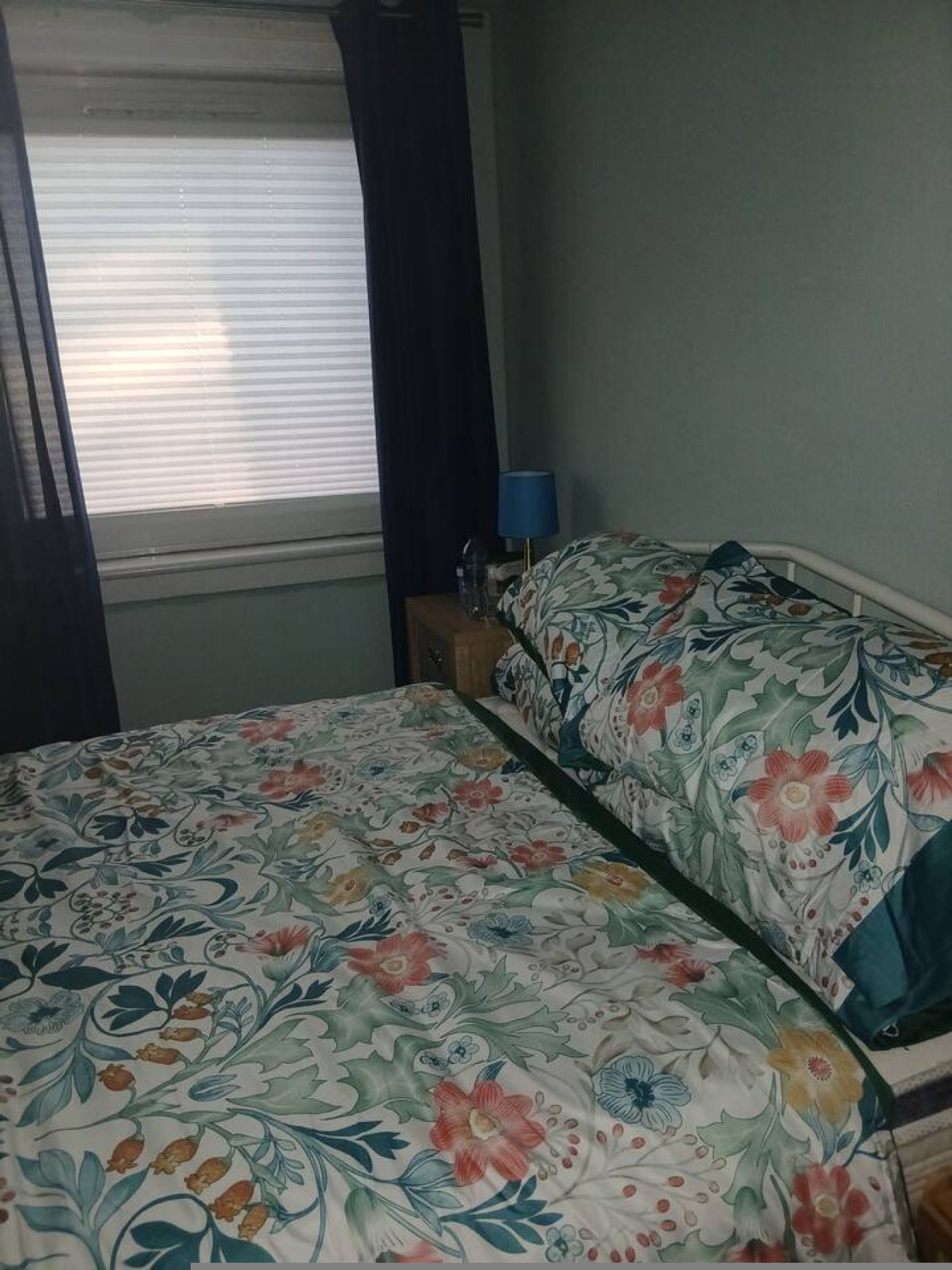 A room with a bed, a window with curtains, a bedside table with a lamp, and a wall with a floral pattern.