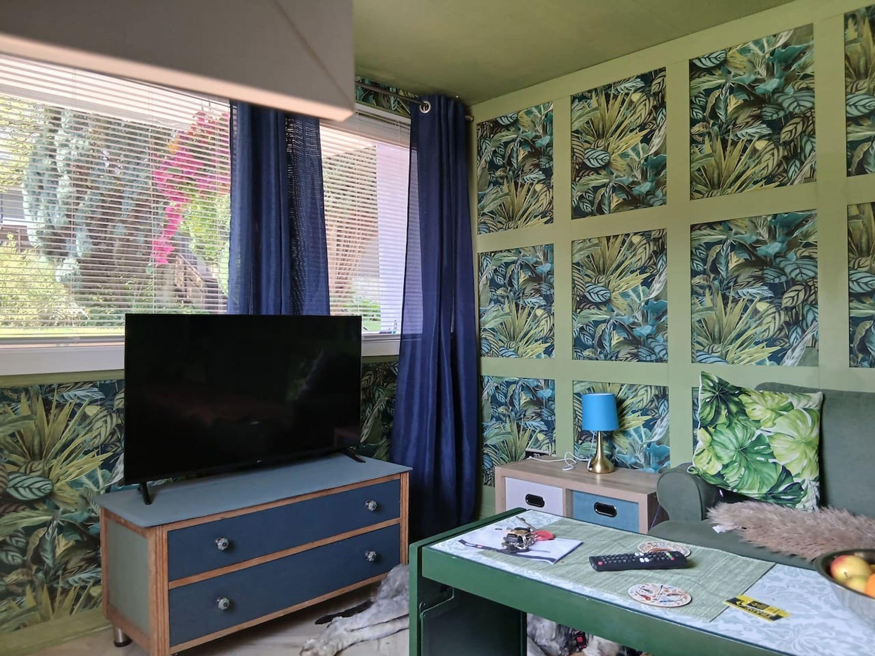 Una habitación con televisor sobre un aparador, un escritorio con lámpara y un diseño de hojas tropicales en la pared.