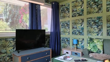 Una habitación con televisor sobre un aparador, un escritorio con lámpara y un diseño de hojas tropicales en la pared.