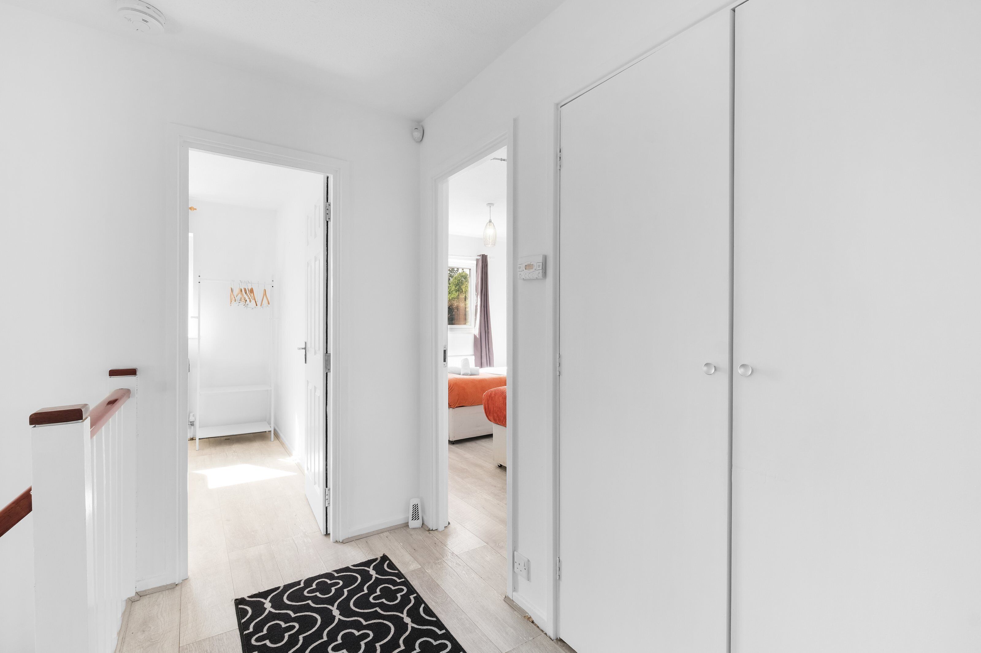 A hallway with white walls, a wooden handrail, and a black patterned rug leading to a bedroom with a bed and a window.