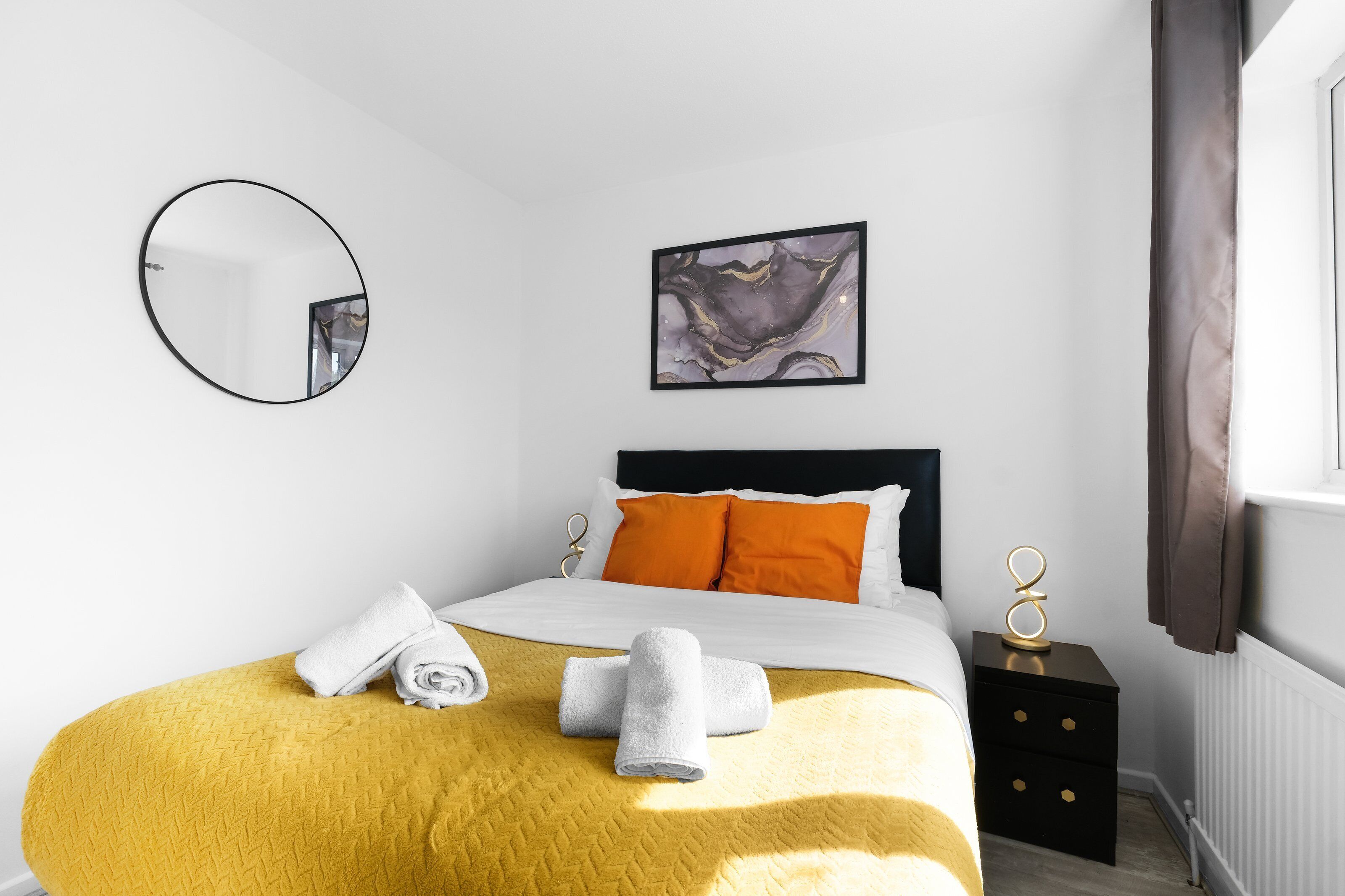 A hotel room with a bed, a round mirror, a painting, and a nightstand.