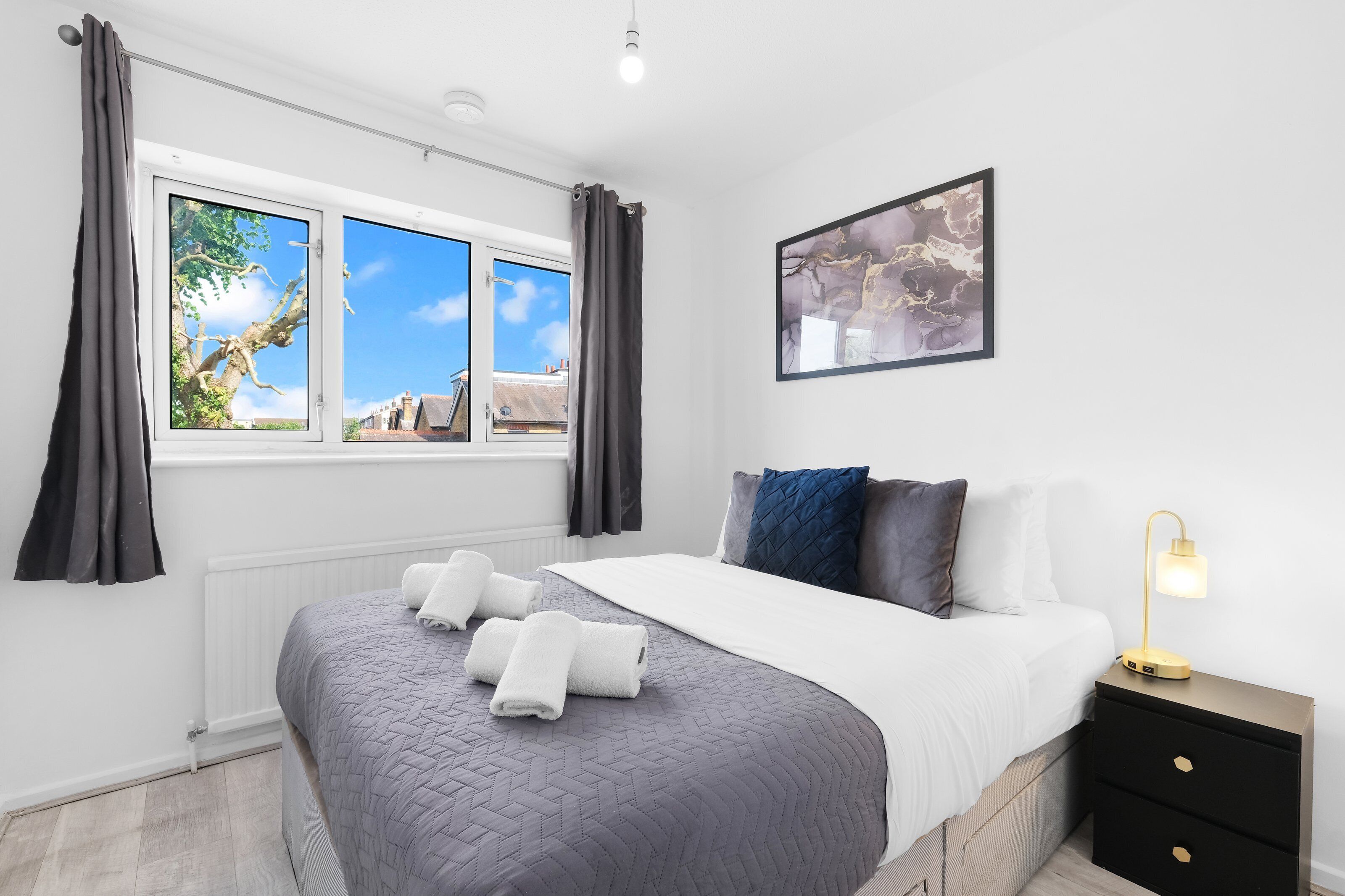 A bedroom with a bed, a nightstand, a lamp, a window with curtains, and a wall-mounted picture.