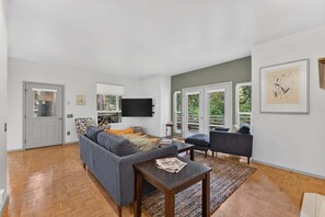 A modern living room with a grey sofa, a coffee table, and a TV mounted on the wall. There are two windows with a view of greenery outside.