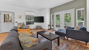 Amplia sala de estar con un sofá gris, un sillón azul y una mesa de centro. Hay un televisor de pantalla plana montado en la pared y una ventana con vistas a árboles.