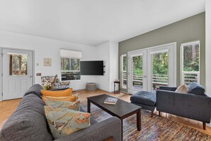 Ein geräumiges Wohnzimmer mit grauem Sofa, blauem Sessel und Couchtisch. An der Wand ist ein Flachbildfernseher angebracht, und ein Fenster bietet Blick auf Bäume.