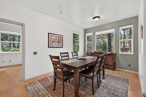 A dining room with a wooden table and chairs, large windows, and a framed picture on the wall.