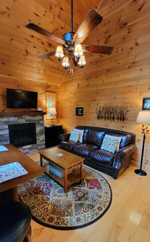 A cozy living room with a fireplace, leather sofa, wooden ceiling, and a television. - Fernwood Hollow- Romantic One Bedroom in Bryson City NC (Bryson City)