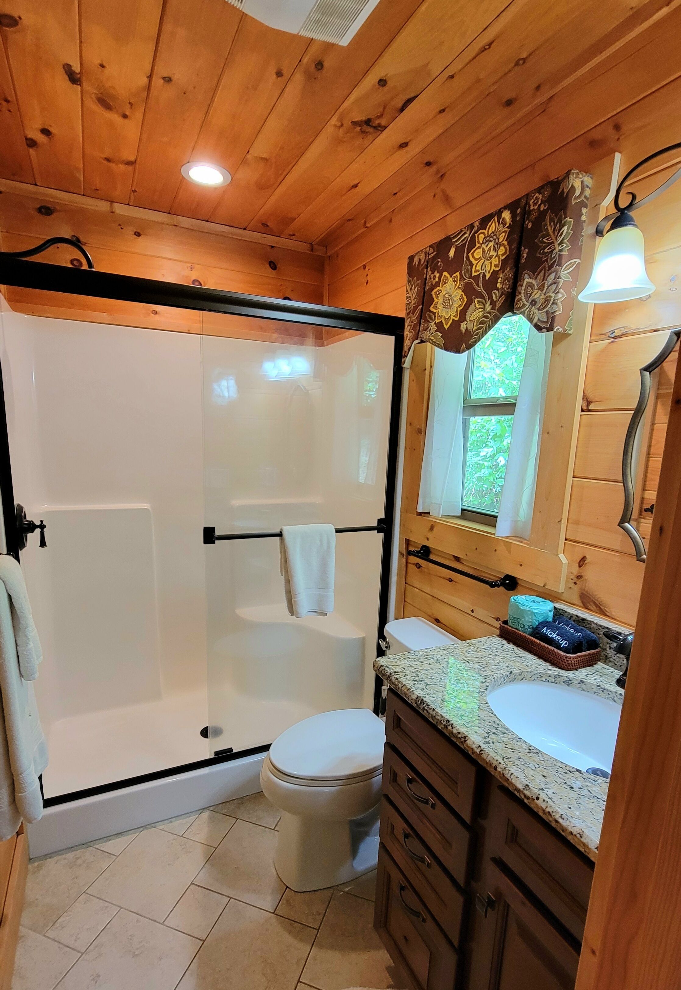 Une salle de bain rustique, avec une cabine en bois, une douche fermée par des parois de verre, des toilettes, un lavabo doté d’un plan de travail en granit et un miroir agrémenté d’un rideau à motifs floraux.