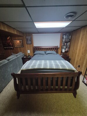 Une pièce avec un lit en bois, un canapé, une table de chevet, une étagère fixée au mur avec des livres, et une fenêtre avec des stores.