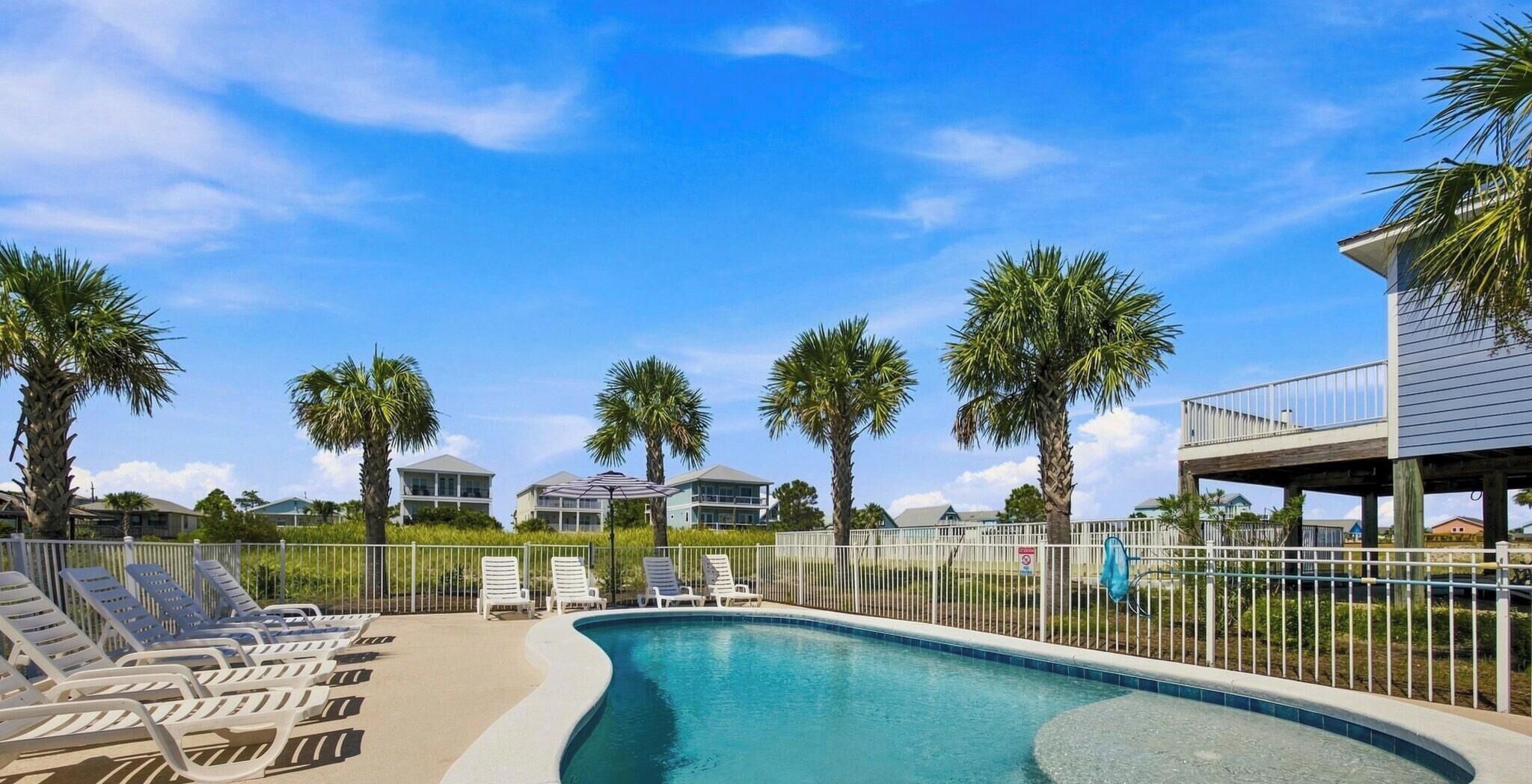 A swimming pool with lounge chairs, palm trees, and houses in the background.
