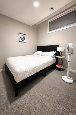 A bedroom with a bed, a bedside table with a lamp, a wall-mounted picture, and a standing fan. - Luxury 2‑bed walkout Suite near CalgaryYYC airport (Calgary)