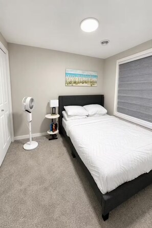 A bedroom with a bed, a standing fan, a nightstand with a lamp, and a painting on the wall. - Luxury 2‑bed walkout Suite near CalgaryYYC airport (Calgary)