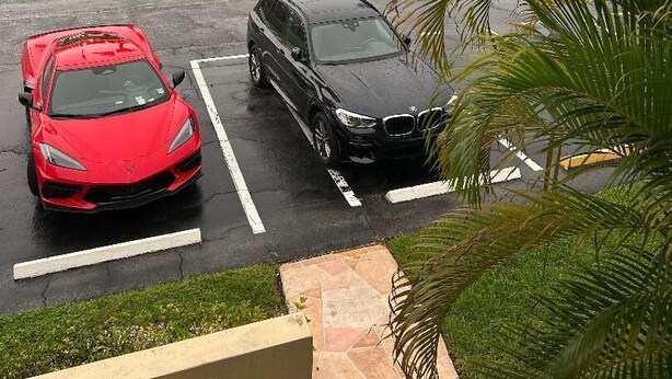 A parking area with two cars, one red and one black, parked on a wet surface with palm trees and a grassy area in the background. - CONDO IN DELRAY BEACH (Delray Beach)