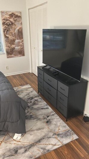 Ein Schlafzimmer mit einem großen Flachbildfernseher auf einem schwarzen Standfuß, einem grauen Sofa und einem Teppich mit Marmoroptik.