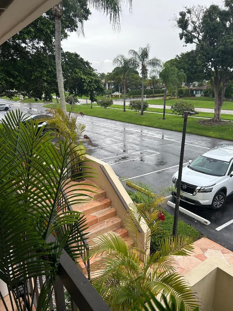 A residential street with a white SUV parked on the side, a staircase leading to a balcony, and lush greenery.