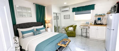 Une chambre à coucher comprenant un lit, une table de chevet, une lampe, une chaise, une petite table avec un livre, et un coin cuisine avec un évier, une cuisinière et un réfrigérateur.