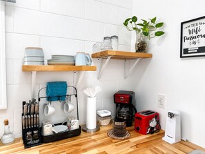 Een keuken met witte betegelde wanden, houten planken, een koffiezetapparaat, een broodrooster en een lijst met een bordje.