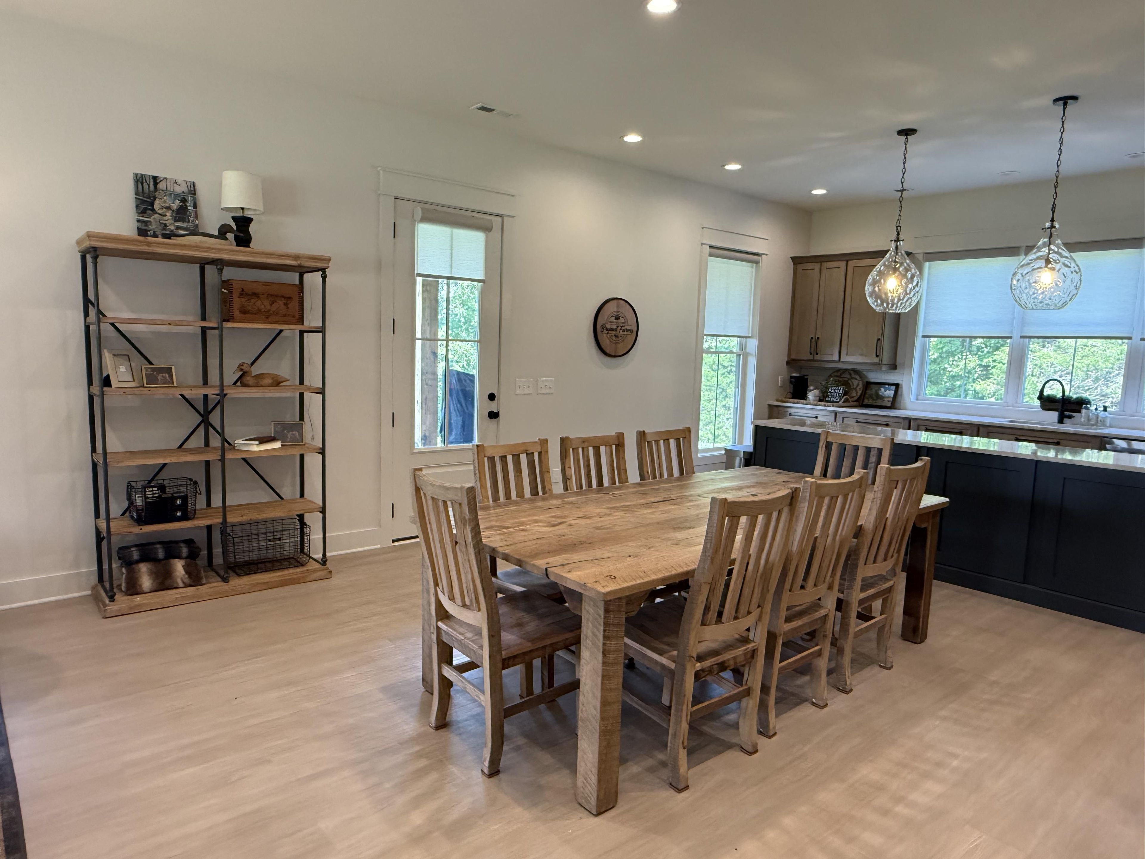 A dining area with a wooden table and chairs, a bookshelf, and a kitchen with a window.