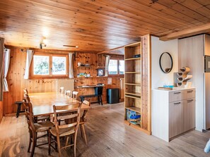 Ein Holzblockhaus-Interieur mit Esstisch, Stühlen, einer Küchenzeile mit Schränken und einem Kühlschrank, sowie einem Fenster mit Vorhängen.