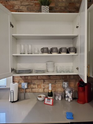 Une cuisine avec un mur en briques, des armoires blanches et divers ustensiles, notamment une bouteille de vin, une bouilloire rouge et une éponge bleue.