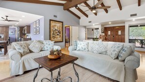 A spacious living room with a high ceiling, wooden beams, a ceiling fan, and a large window offering a view of the outdoors. - West Sedona 3BR retreat with game room, patio, and fenced yard. (Sedona)