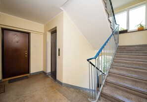 A staircase with a blue handrail leading to an upper floor, a door with a number plate, and a wall-mounted light switch. - Rondo ONZ Homely Apartment (Warszawa)