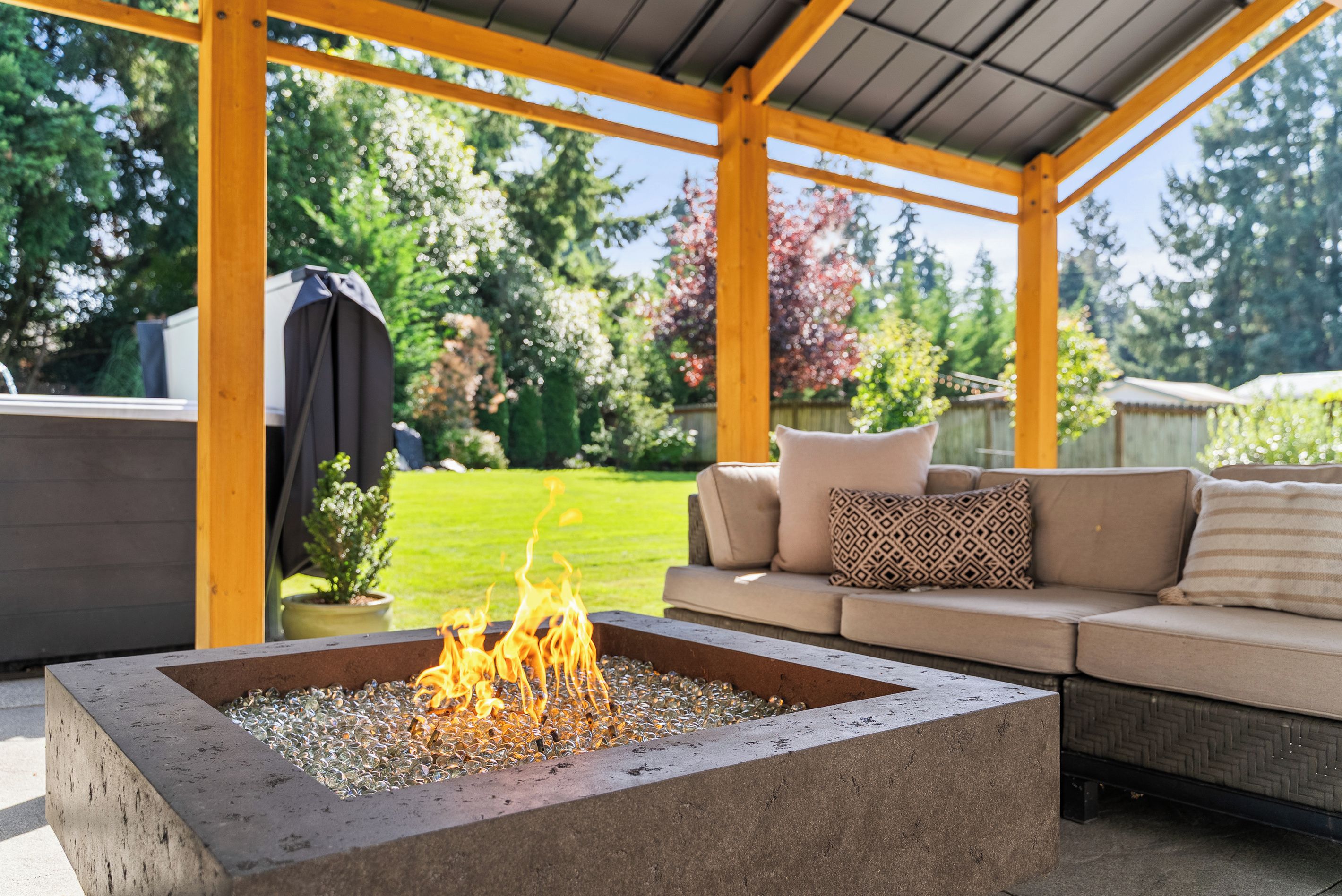 Un espace extérieur aménagé, partiellement abrité, avec un foyer au centre, entouré d’une pergola en bois et d’une pelouse.