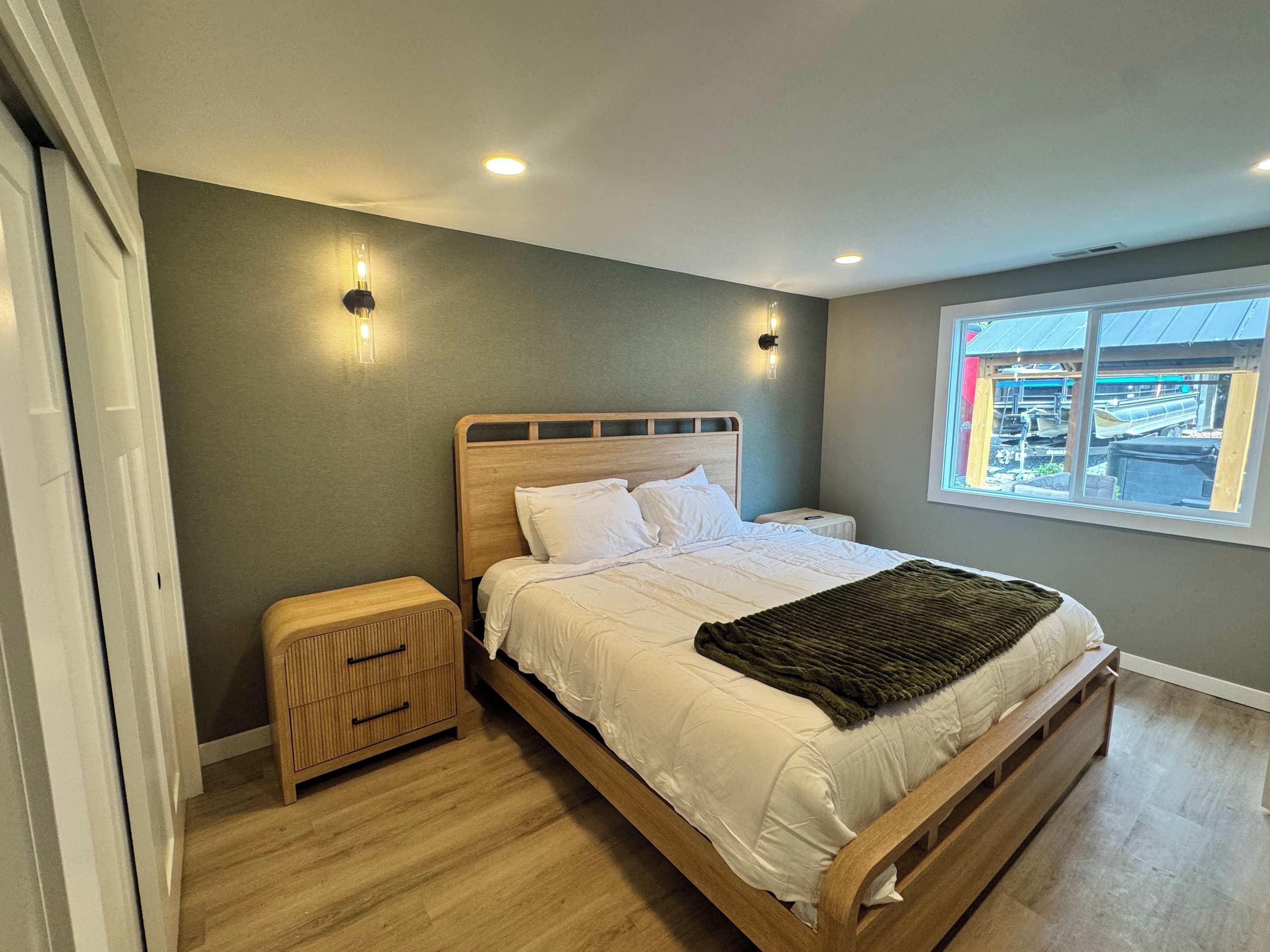 A bedroom with a wooden bed, bedside tables, a window with a view, and wall-mounted lighting.