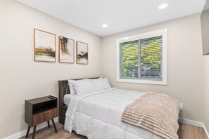 A bedroom with a bed, a nightstand, a window with blinds, and three framed pictures on the wall. - The Terrace | Grason Properties (Puyallup)