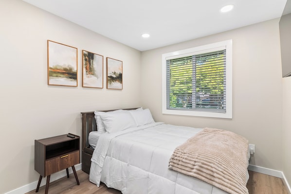A bedroom with a bed, a nightstand, a window with blinds, and three framed pictures on the wall.