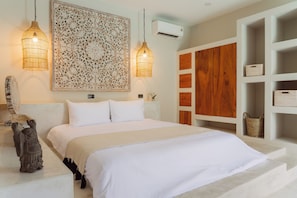A neatly made bed with a white comforter and pillows, two hanging light fixtures, a decorative wall piece, and a wooden panel wall unit with storage baskets.