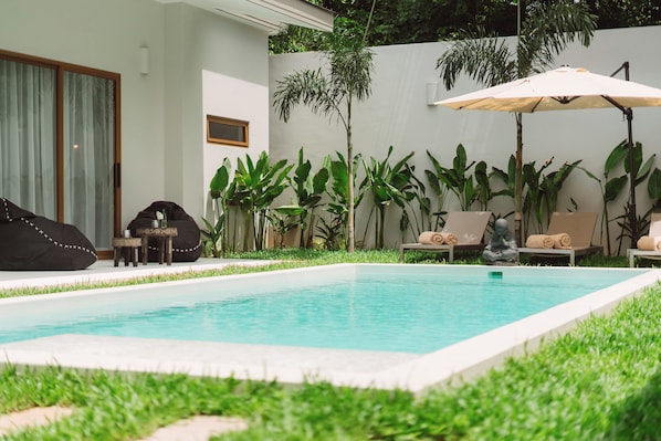 A swimming pool with lounge chairs and a statue, surrounded by lush greenery and a white wall.
