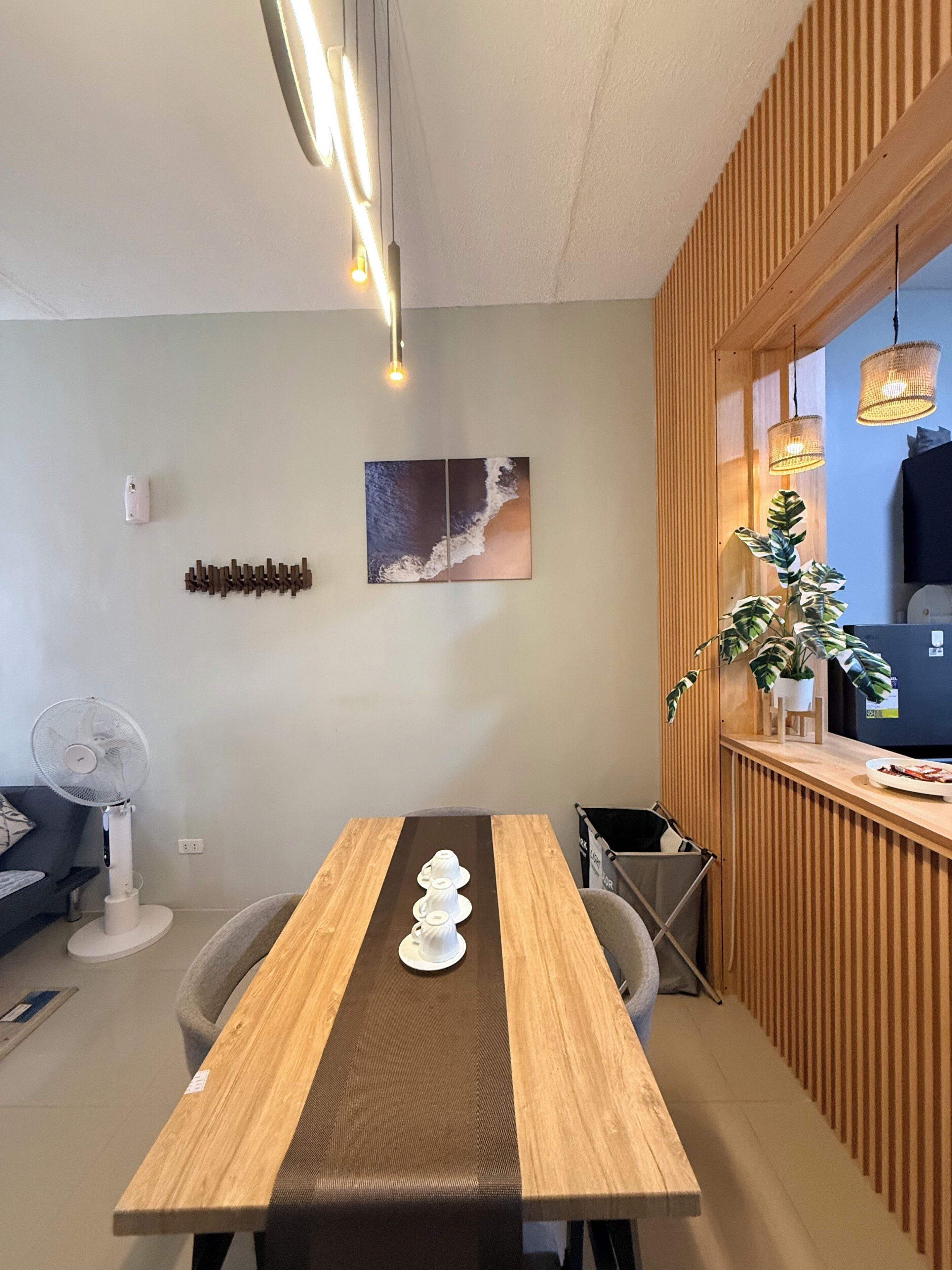 Ein moderner Essbereich mit einem Holztisch, Stühlen, einem Wandventilator, einem dekorativen Wandbild und einem Blumentopf.