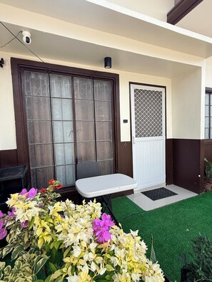 A covered outdoor seating area with a table and chairs, a door with a security camera, and a small garden with various plants.