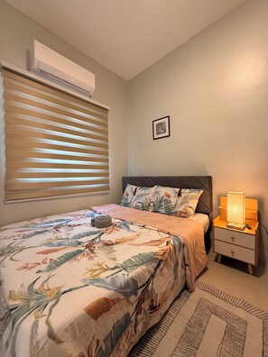 A bedroom with a bed, bedside table, nightstand, window with blinds, and a wall-mounted air conditioner.