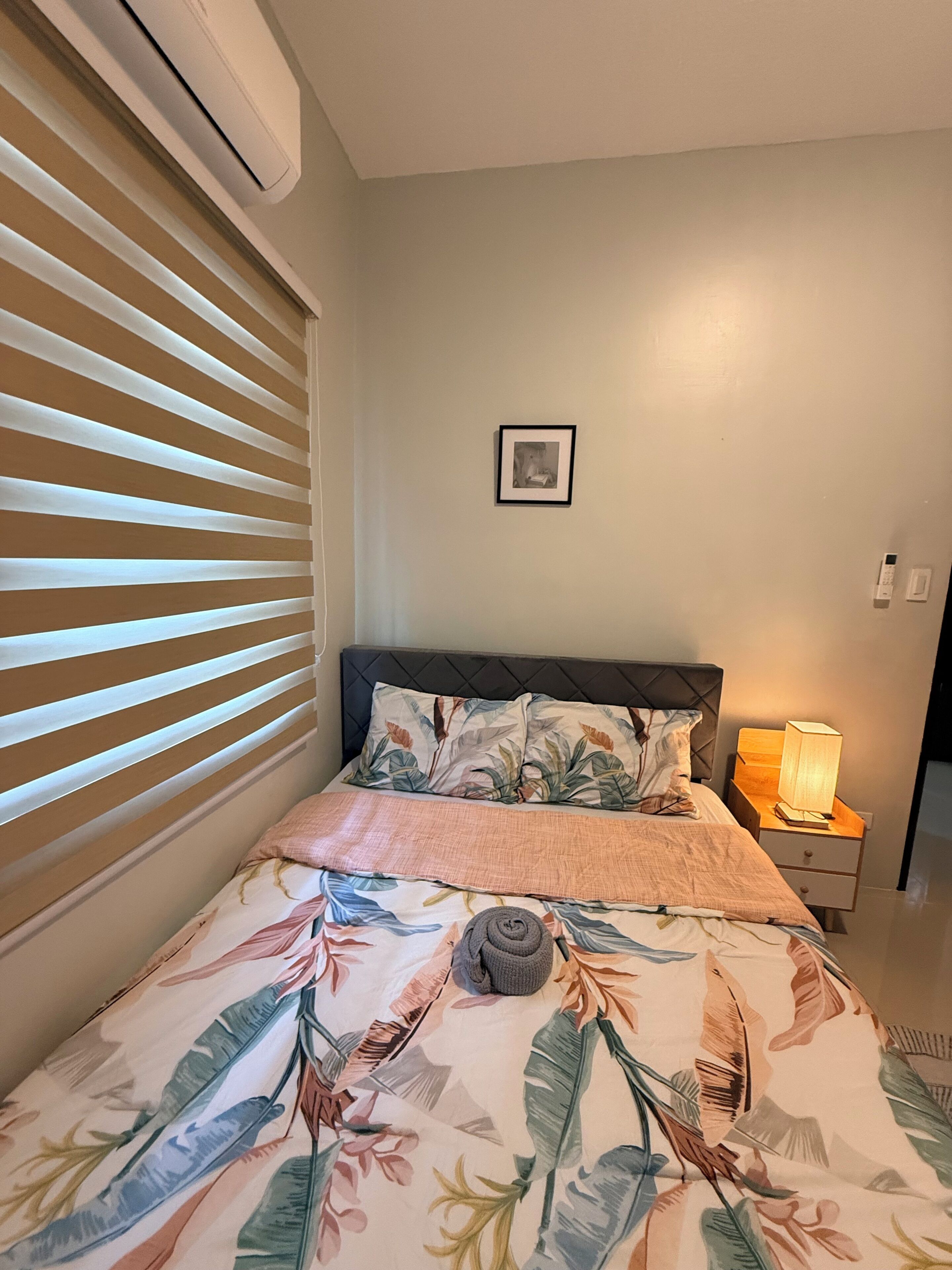 A bedroom with a bed, a nightstand with a lamp, a framed picture on the wall, and a window with blinds.