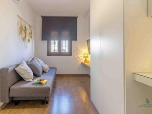 Ein schmales Zimmer mit einer grauen Couch, einem kleinen Fenster und einer Wandlampe.