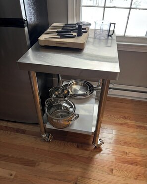 Una cocina con un refrigerador de acero inoxidable, una tabla de cortar de madera con cuchillos, una jarra de vidrio y una olla de metal sobre un carrito metálico.