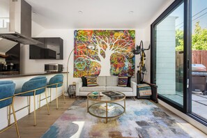 Ein modernes Wohnzimmer mit einer Couch, einem Glastisch, einem farbenfrohen abstrakten Gemälde, einem Fahrrad und einer Glasschiebetür nach draußen.