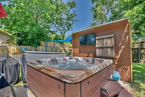 A hot tub in a backyard with a wooden deck, a covered seating area, and a small building with a window.