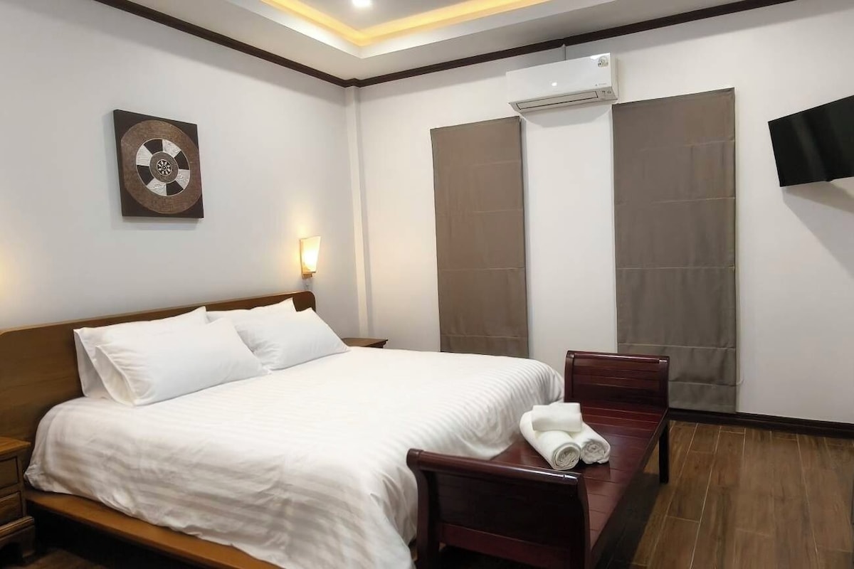 A hotel room with a bed, bedside table, wall-mounted TV, and a large window with curtains.