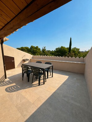 Un patio exterior con una mesa y sillas, una pérgola de madera, y vistas a árboles y un edificio al fondo.