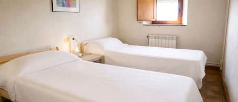 A room with two single beds, a nightstand with a lamp, a radiator, and a window with blinds.
