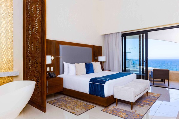 A modern hotel room with a large bed, a view of the ocean, and a balcony with a chair. - Escape to a beautiful two-bedroom oceanfront resort in Cabo. (San José del Cabo)