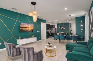A modern living room with a dining area, kitchen, and a large painting on the wall. - Themed 7BR by Disney | Heated Pool + Game Room (Davenport)