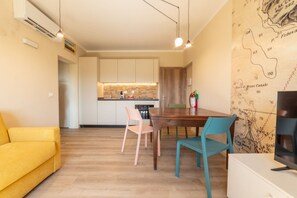 A modern living room with a wooden floor, a yellow sofa, a wooden dining table with chairs, and a kitchen in the background. - APPARTAMENTI IL SENTIERO DEL GRANITO - GRANITO BILO 1 DX (Campo nell'Elba)