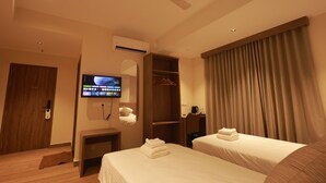 A hotel room with two beds, a television, an air conditioning unit, a ceiling fan, a wardrobe, and a window with curtains.